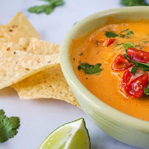 creamy vegan cashew queso in a bowl with tomatoes, cilantro, chips, chiles, and limes.
