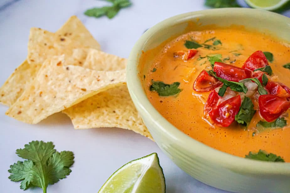 creamy vegan cashew queso in a bowl with tomatoes, cilantro, chips, chiles, and limes.