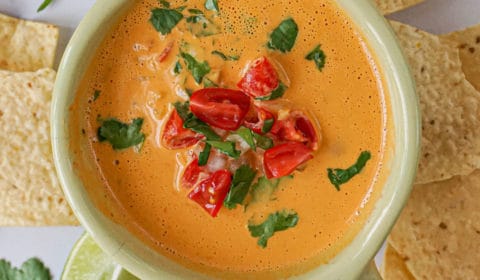 creamy vegan cashew queso in a bowl with tomatoes, cilantro, chips, chiles, and limes.