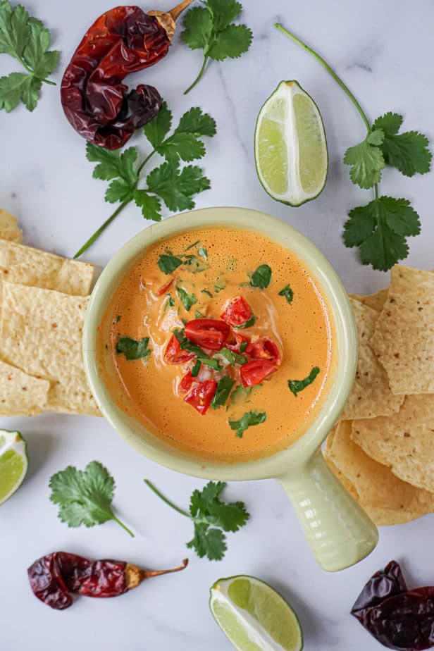 creamy vegan cashew queso in a bowl with tomatoes, cilantro, chiles, lime, and chips.