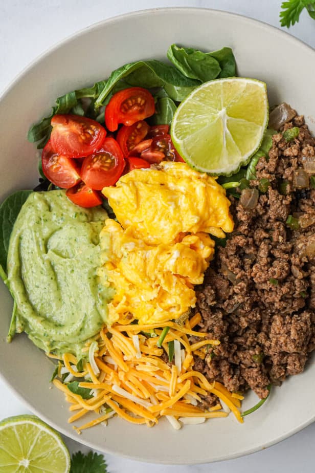 High Protein Low Carb Cookbook recipe a spicy southwest breakfast bowl with eggs, ground beef, tomato, lime, avocado sauce, and greens.